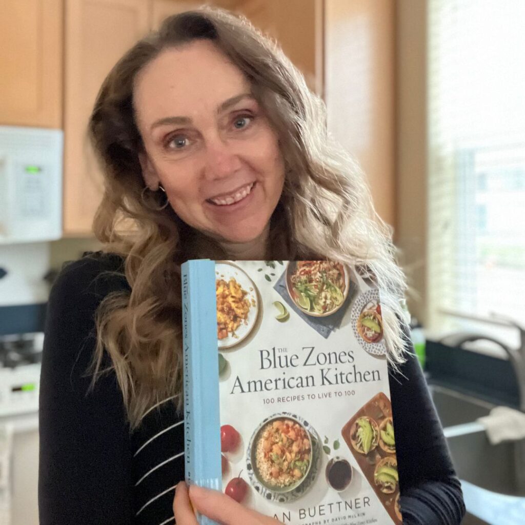 A photo of Gorgeous Wellth founder Lori Osterberg with the cookbook The Blue Zones American Kitchen as a part of her Cookbook Club
